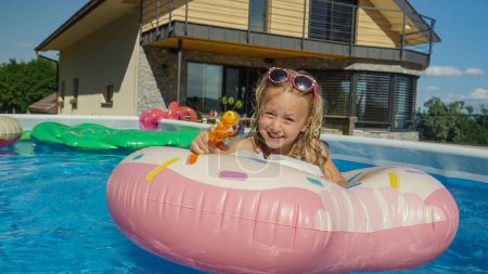 Foto de CLOSE UP: Adorable girl lying on a floatie pointing a water gun towards camera. Cheerful little girl floating in backyard swimming pool and having fun with water blaster pointing in camera direction. - Imagen libre de derechos