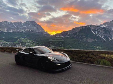 Téléchargez les photos : Sports car driving above alpine mountain town under gorgeous sunset in Dolomites. Glowing colorful skyscape above high mountain peaks and town in the valley. Picturesque moment at the end of the day. - en image libre de droit