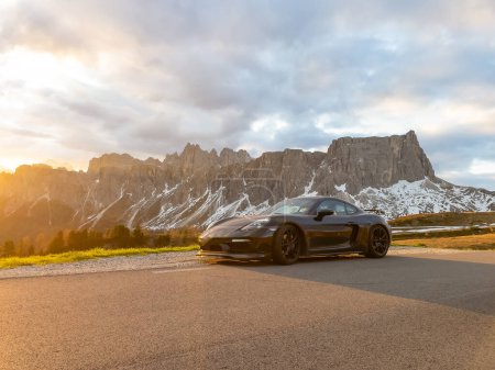 Téléchargez les photos : GIAU PASS, DOLOMITES, ITALY, MAY 2022: Black car in golden light with amazing mountain backdrop in Dolomites. Sports car at roadside of winding mountain road with gorgeous alpine peaks in background. - en image libre de droit