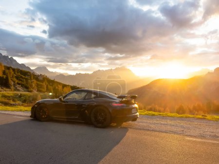 Téléchargez les photos : GIAU PASS, DOLOMITES, ITALY, MAY 2022: Picturesque sunset in the alpine mountains with a black sports car at roadside. Burning clouds above mountain peaks and last rays of sun peeking through. - en image libre de droit