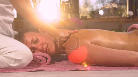 Photo for Relaxed young woman receiving a therapeutic back massage in the morning. Sun rays shinning through female hands while gently massaging back of a young lady. Massage therapist using body oil. - Royalty Free Image