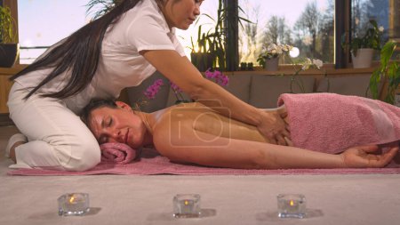 Foto de Young female person relaxing while receiving therapeutic back massage. Sun rays shinning through female hands while gently massaging back of a young lady. Spa treatment at the end of the day - Imagen libre de derechos