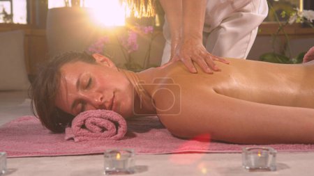 Téléchargez les photos : Young lady relaxing while having a pleasant neck and shoulder massage. Golden sun rays shinning through hands of a female therapist while gently massaging shoulder and neck of a young lady. - en image libre de droit