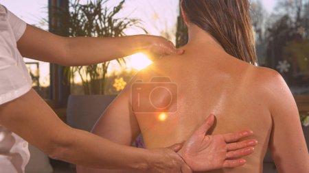 Foto de Female therapist massaging and relieving young woman's back muscles. Relaxing therapeutic massage in the embrace of golden sunlight. View of female hands performing muscle knot relief massage - Imagen libre de derechos