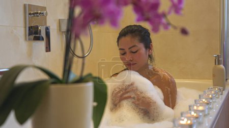 Téléchargez les photos : Belle jeune femme asiatique appréciant prendre un bain moussant dans une ambiance relaxante. Détente et soins spa dans la salle de bain à la maison pour la fin de la journée. Dame se traiter avec bain plein de mousse. - en image libre de droit