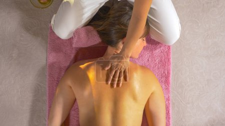Photo for Therapeutic masseuse massaging upper back and shoulder of a young lady. Female massage therapist using massage oil for deep muscle relaxation. Wellness treatment for back muscle relief. - Royalty Free Image