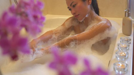 Téléchargez les photos : Pretty young lady relaxing and washing herself in bath full of foam. Beautiful Philippine woman enjoying at spa treatment in home bathroom while taking a bubble bath in a relaxing ambience. - en image libre de droit