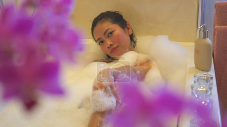 Téléchargez les photos : Beautiful Philippine woman enjoying while taking a nice bubble bath. Young female person relaxing while lying in bath full of foam and looking into camera. Beauty treatment in home bathroom. - en image libre de droit