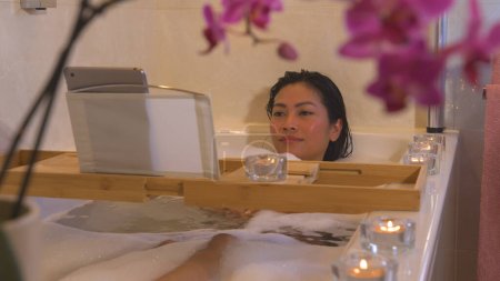 Téléchargez les photos : Relaxed young woman lying in bubble bath and watching an online movie. Beautiful Philippine lady relaxing in bathtub after a busy day and watching her favourite series on digital tablet. - en image libre de droit