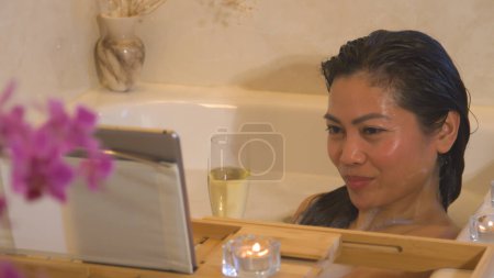 Téléchargez les photos : Woman in foam bath holding champagne glass during online romantic date. Beautiful Philippine woman in relaxing bubble bath, smiling and flirting with husband via video call on digital tablet - en image libre de droit
