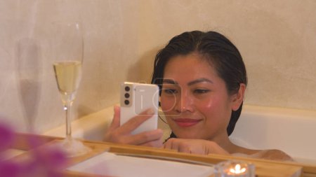 Téléchargez les photos : Young female in bubble bath taking photos of full glass of champagne. A beautiful Philippine woman pampering herself with luxurious spa treatment and making pictures to post on social media - en image libre de droit