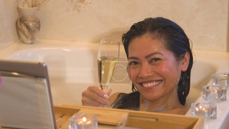 Téléchargez les photos : Pretty Philippine lady enjoys in bubble bath holding glass of champagne. A beautiful woman treating herself with luxurious pampering while relaxing in bathtub full of foam after a busy day. - en image libre de droit