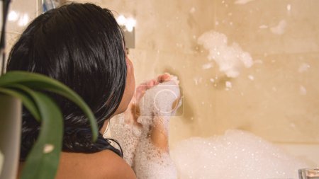 Téléchargez les photos : Rear view shot of playful dark haired woman blowing foam in bubble bath. Female person chilling and enjoying in bathtub full of foam surrounded with candles. Relaxation after a long busy day - en image libre de droit