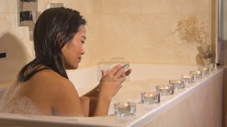 Téléchargez les photos : Beautiful young woman texting on phone while having a bubble bath. Smiling Philippine lady responding on text message on smartphone while taking a foamy bath in nice ambience with candles - en image libre de droit