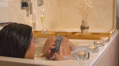 Téléchargez les photos : Rear view shot of young lady with cell phone while soaking in the bath. Dark haired female person enjoying and relaxing in bubble bath while using smartphone for scrolling over social media. - en image libre de droit