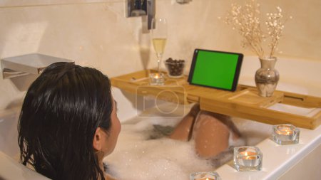 Téléchargez les photos : Relaxed young lady chilling in bubble bath and watching online video. Beautiful woman relaxing at home bathroom and taking time for herself. Home wellness for stress relief and body care. - en image libre de droit