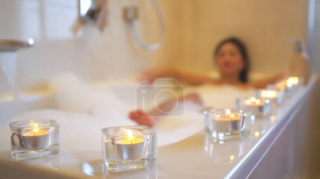 Foto de Alineado velas brillantes para relajarse ambiente mientras toma un baño de burbujas agradable. Hermosa mujer asiática joven tratando a sí misma con spa y relajarse mientras se enfría en un bonito baño lleno de espuma burbujeante. - Imagen libre de derechos