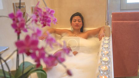 Téléchargez les photos : Belle fleur d'orchidée pourpre fleurie avec jeune dame relaxant dans un bain pétillant en arrière-plan. Belle jeune femme relaxante dans la baignoire. Ambiance agréable et agréable pour une expérience de soins spa. - en image libre de droit