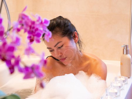 Téléchargez les photos : Pretty young Asian lady having a bubbly bath and washing her body. Beautiful Philippine lady body washing herself with bath foam. Time for personal hygiene in relaxing home ambience. - en image libre de droit