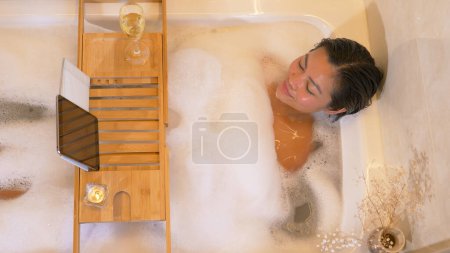 Téléchargez les photos : Pretty young lady having relaxation time in bath and watching a movie. Young lady enjoying a nice bubble bath. Beautiful Philippine woman watching comedy movie while soaking in foamy bath. - en image libre de droit