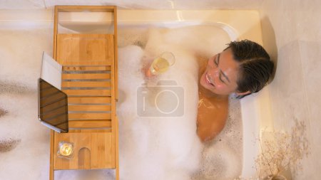 Téléchargez les photos : Gorgeous lady in bubble bath drinking champagne and having video chat. Young woman enjoying a nice bubble bath. Relaxation time with online video call and wellness treatment for the evening. - en image libre de droit