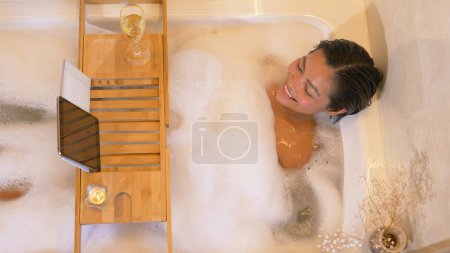 Téléchargez les photos : Attractive Philippine woman in bubble bath and talking via video chat. Young lady enjoying a nice bubble bath. Relaxation time with online video call and wellness treatment at the end of day - en image libre de droit