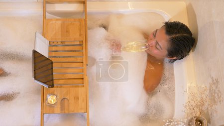 Téléchargez les photos : Beautiful lady in bubble bath drinking champagne and having video chat. Young woman enjoying a nice bubble bath. Relaxation time with online video call and wellness treatment for the evening - en image libre de droit