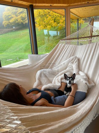 Foto de Rear view of beautiful young woman chilling in hammock on rainy autumn day. Pretty lady relaxing with miniature pinscher while looking at autumn colored garden trees through big living room windows. - Imagen libre de derechos