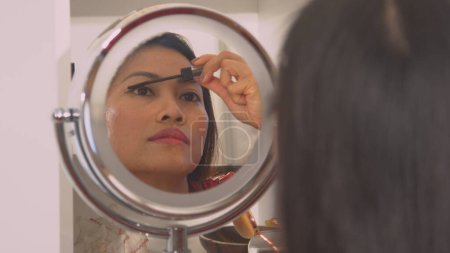 Téléchargez les photos : Pretty Philippine lady in mirror reflection applying black mascara. Young woman putting on make up for an evening date in front of a mirror. Female person taking care of her appearance. - en image libre de droit