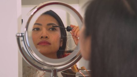 Téléchargez les photos : Beautiful Philippine woman in mirror reflection applying black mascara. Young lady putting on make up for an evening date in front of a mirror. Female person taking care of her appearance. - en image libre de droit