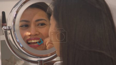Téléchargez les photos : Mirror reflection of a woman flossing her teeth for tartar prevention. Beautiful young lady using dental floss toothpick for taking care of dental hygiene. Morning routine for a nice smile. - en image libre de droit
