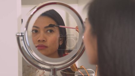 Téléchargez les photos : Young woman correcting eyebrow shape with brush in mirror reflection. Pretty Philippine lady beautifying and shaping her eyebrows with brow comb. Female person perfecting her face make up. - en image libre de droit