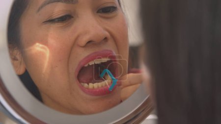 Téléchargez les photos : View of beautiful woman in cosmetic mirror flossing her white teeth. Pretty young lady using dental floss toothpick for taking care of dental hygiene. Morning routine to keep a nice smile. - en image libre de droit