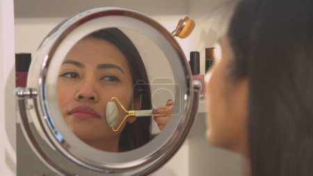 Téléchargez les photos : Young woman using face roller for self massaging her beautiful face. Pretty Philippine lady at rejuvenating face treatment. Mirror reflection of female person taking care of her appearance. - en image libre de droit