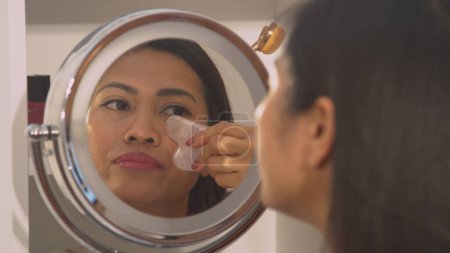 Téléchargez les photos : Pretty Philippine woman using jade scraper for self massaging her face. Mirror reflection of beautiful young lady using gua sha scraper for anti wrinkle and face slimming beauty treatment. - en image libre de droit