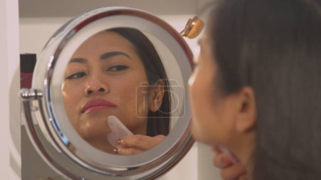 Foto de Reflection of beautiful woman in cosmetic mirror using gua sha scraper. Young Philippine lady at rejuvenating beauty treatment using jade roller for skin firmness and face lifting massage. - Imagen libre de derechos