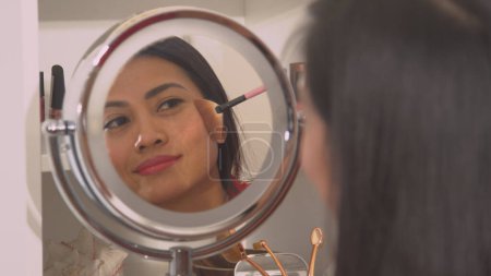 Téléchargez les photos : Attractive Philippine woman applying bronzer to beautify her face. Mirror reflection of a pretty lady using brush to apply face blush. Female person taking care of her fresh look. - en image libre de droit