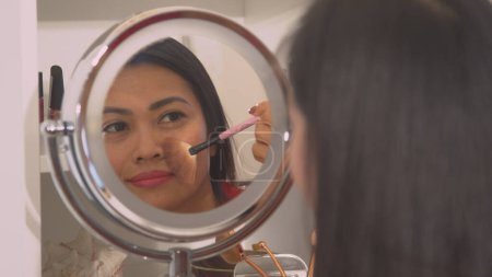 Téléchargez les photos : Attractive young Asian woman applying powder bronzer to beautify her face. Mirror reflection of a pretty lady using brush to apply face blush. Female person taking care of her fresh look. - en image libre de droit