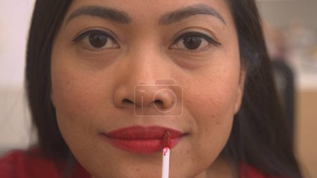Téléchargez les photos : Attractive Philippine woman applying vivid red lip gloss to her lips. Pretty young lady using colourful and shiny lip product. Female person perfecting her look, finishing make up treatment. - en image libre de droit