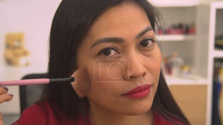 Téléchargez les photos : Young Philippine lady using brush for applying bronzer to her face. Mirror reflection of pretty woman applying face blush to beautify her face. Female person taking care of her fresh look. - en image libre de droit