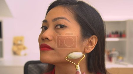 Photo for Close up view of young lady self massaging her face with jade scraper. Beautiful Asian woman using gua sha scraper for rejuvenating anti-aging massage and facial muscles relief treatment. - Royalty Free Image