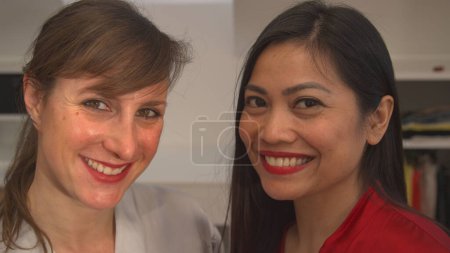 Téléchargez les photos : PORTRAIT: Smiling young ladies ready for a night out after finishing their glamourous make up. Asian and Caucasian woman applying facial beauty products. Two females having fun while doing makeover. - en image libre de droit