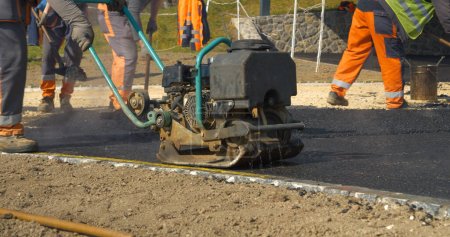 Photo for APRIL 2022, LJUBLJANA, SLOVENIA: Construction worker using vibration compactor for laying asphalt surface at yard. Working men in beautiful morning light. Men in uniform asphalt paving the driveway. - Royalty Free Image
