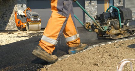 Photo for APRIL 2022, LJUBLJANA, SLOVENIA: Rear view of worker using vibration compactor for smoothing asphalt surface. Working men in beautiful morning light. Men in uniform asphalt paving the driveway. - Royalty Free Image