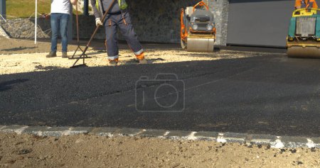 Téléchargez les photos : CLOSE UP: Partially asphalted and levelled fresh asphalt surface on the driveway. Man at work asphalt paving the driveway in beautiful morning light. Man in uniform working on asphalting the yard. - en image libre de droit