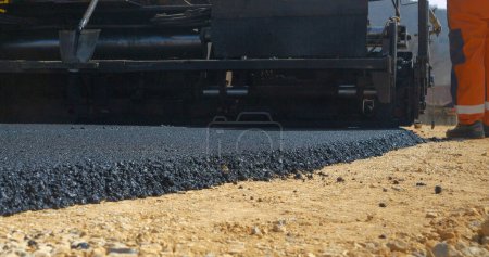 Photo for CLOSE UP: View from back of paver machinery leaving behind freshly laid asphalt. Heavy operating construction machinery dumping and spreading fresh blacktop surface at driveway in nice morning light. - Royalty Free Image