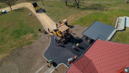 Photo for AERIAL: Group of construction workers laying asphalt concrete on yard and road. Men in uniform asphalt paving driveway. Builders preparing terrain for finishing paving the roadway in morning light. - Royalty Free Image