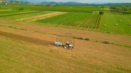 Foto de AERIAL: White tractor with slurry spreader fertilizing arable land in autumn. Farm worker spreading manure on the field with modern farming machinery on a sunny day. Agricultural tasks in fall season. - Imagen libre de derechos