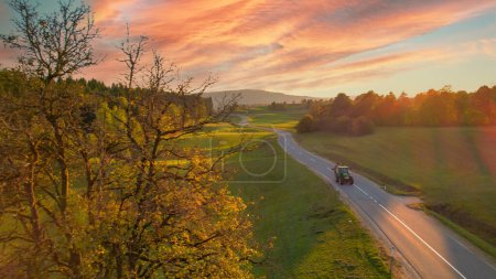 Photo for AERIAL: Tractor returning and driving on paved road after tedding mowed grass. Farm vehicle with attached agricultural machinery driving along asphalt country road in beautiful golden autumn sunlight. - Royalty Free Image