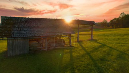 Foto de AERIAL: La luz del sol de otoño se asoma sobre el borde de un pajar tradicional. Antiguo edificio de la granja para el secado y almacenamiento de heno de pie en medio de un hermoso prado verde con luz dorada. - Imagen libre de derechos